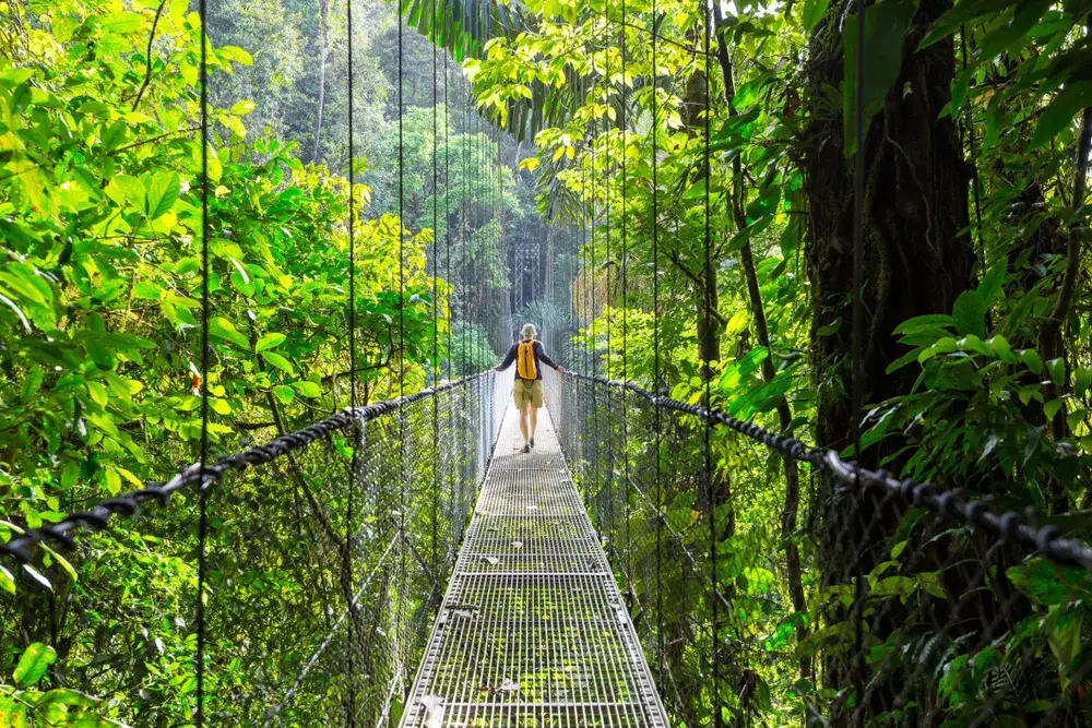 For a piece titled Is Costa Rica Safe to Visit, a fit blonde woman with a backpack walking along a suspension bridge in a rainforest