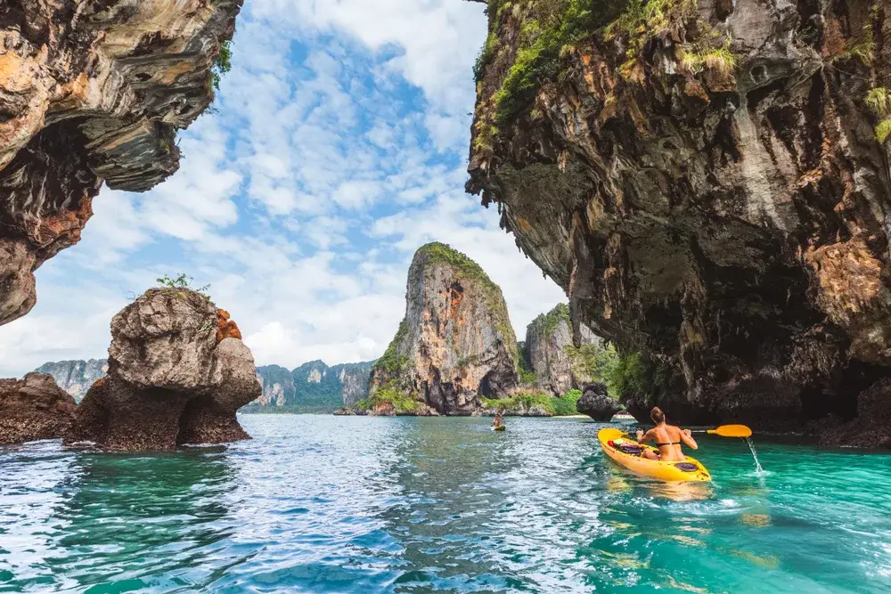 Tan brunette woman paddling a yellow kayak through a rock formation in the Krabi province, one of the must-visit places in Thailand