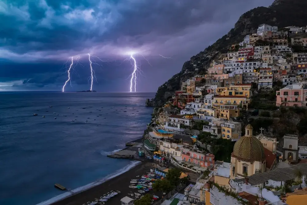 Lightning striking the ocean in the distance as seen from the POV of a person standing on a rooftop with dark storm clouds overhead during the worst time to visit Positano