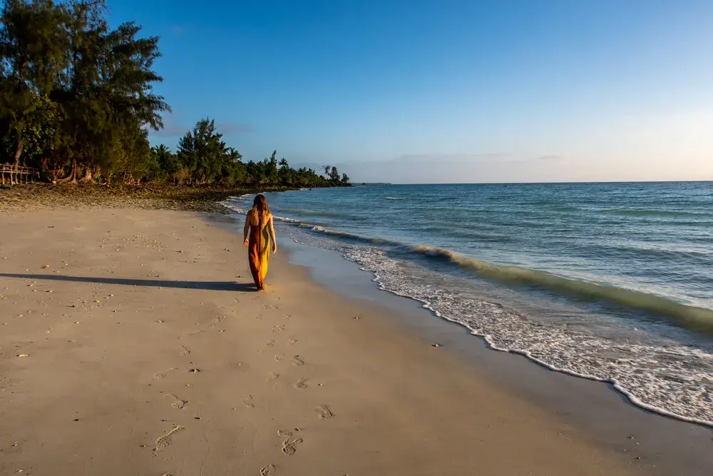 Woman in a long orange dress walking along a beach next to the ocean with clear skies during the least busy time to visit Tanzania
