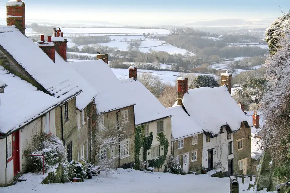 Brick homes along a sloping road in Gold Hill Shaftesbury with snow covering the roofs, landscaping, and roads during the winter, the cheapest time to visit England