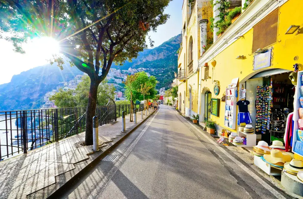 Empty street with semi-open shops selling colorful hats and clothing with the sun rising over the mountains on the left side during the least busy time to visit Positano