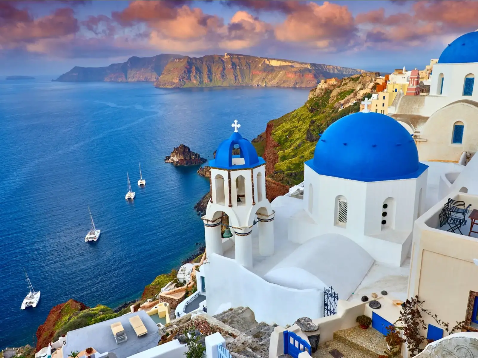 A view from Fira Town with its peculiar houses and vibrant rounded roofs at Santorini Island, one of the best islands in Greece to visit, overlooking four sailing boats in a vast blue sea