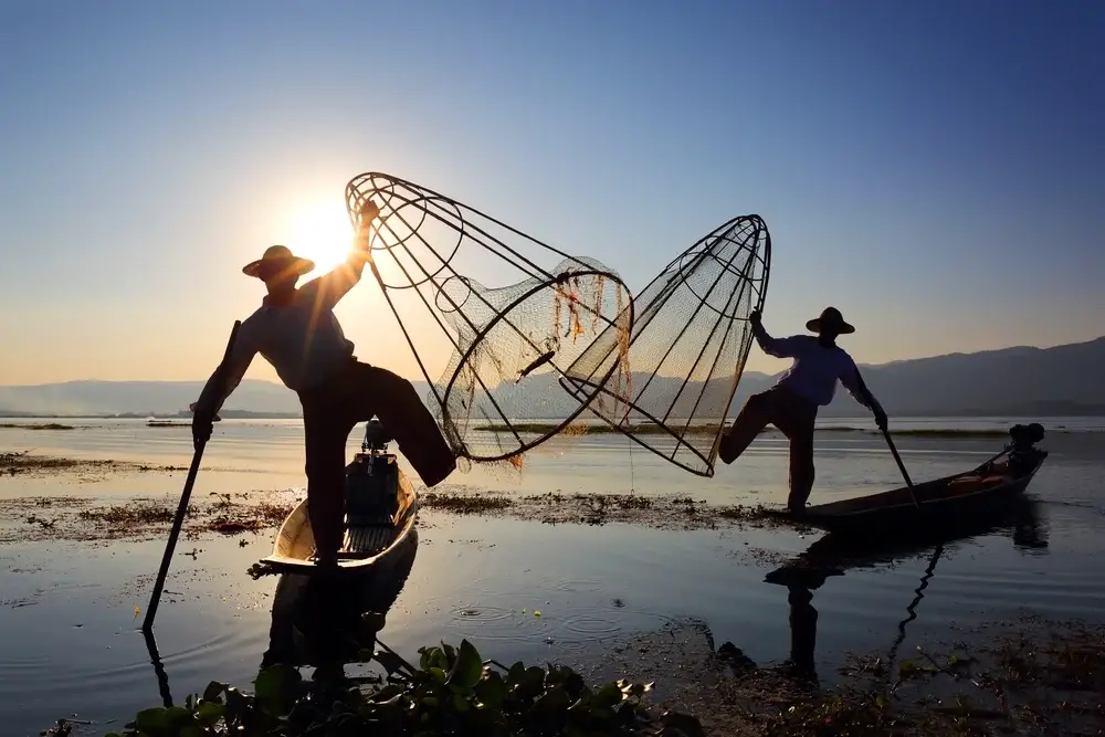 Burmese men in Myanmar pictured doing their one-leg rowing style while holding net in the other hand at dusk