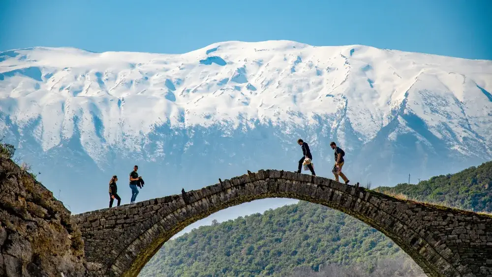 People crossing the Old Bridge at the thermal baths in Albania in front of snowy mountains during the cheapest time to visit Albania