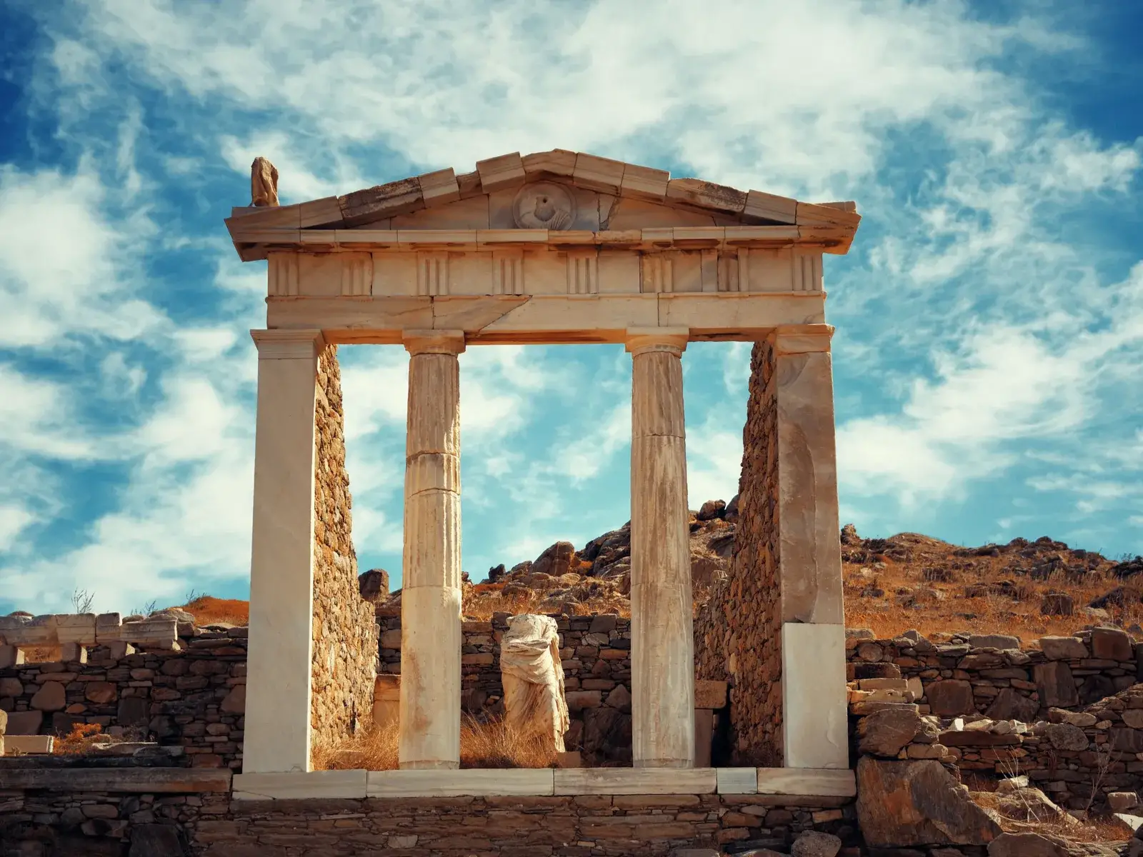 Historical ruins of a large hall with two columns and the lower portion of a broken statue seen at Delos Island as one of the best islands in Greece to visit