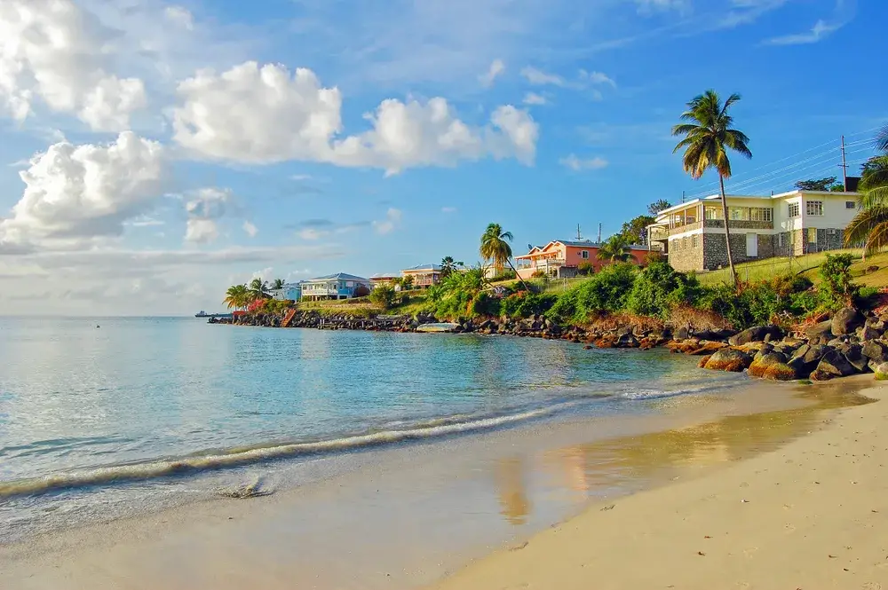Still water of Grand Anse Beach pictured with waves lightly lapping the tan sand and gorgeous homes overlooking the bay with blue skies overhead