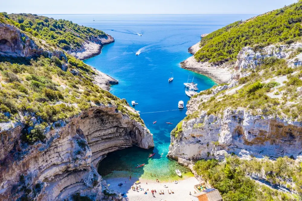 Rugged cliffs towering over a small sandy bay in Vis, one of the best places to visit in Croatia, with a number of tourists on the sand below